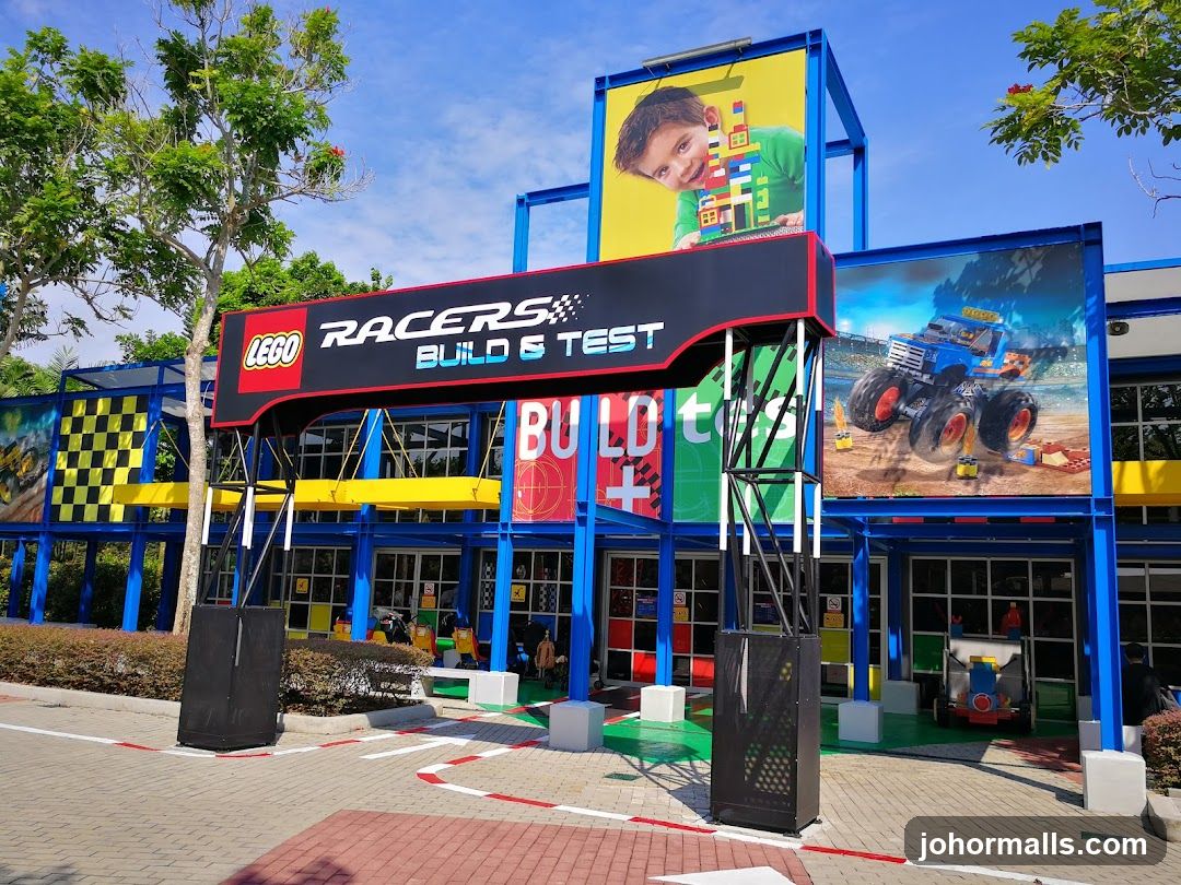 Exterior view of a brightly colored building facade with blue, yellow, and red accents. Large sign reads 'LEGO RACERS BUILD & TEST' above the entrance. Prominent advertising banners feature LEGO characters and monster trucks. The ground has painted racing lines. Trees and clear sky in background.