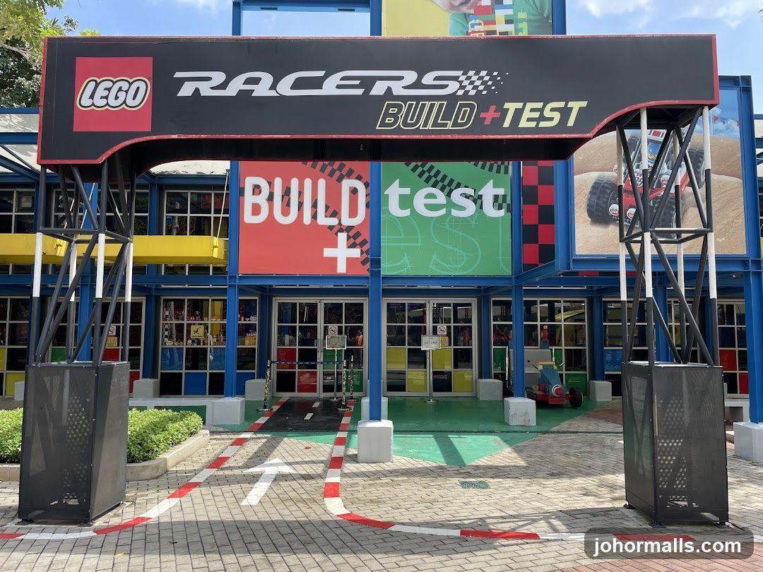 Exterior view of a Lego-themed storefront with a large black archway sign displaying 'LEGO RACERS BUILD+TEST'. The building facade is blue with colorful panels showing 'BUILD + test' in red and green. A paved entrance area features a race track-style path with red and white markings leading to glass doors.