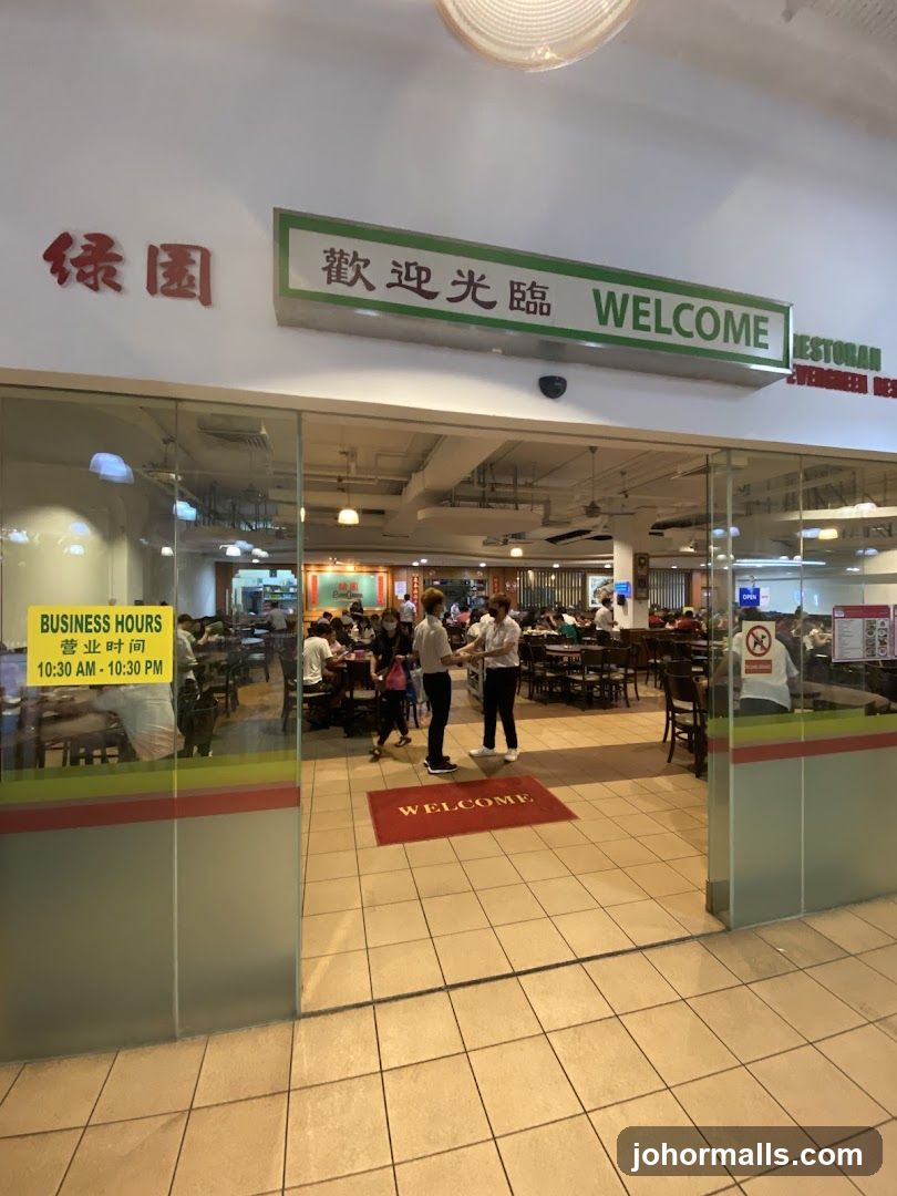 Glass storefront with a green and white sign above reading '歡迎光臨 WELCOME' and red Chinese characters '绿园' to the left. A yellow business hours sticker is on the glass door. Inside, patrons are seated at tables; staff in white shirts are present. A red 'WELCOME' mat lies on tiled floor. The interior shows multiple dining areas with wooden furniture and hanging lights.