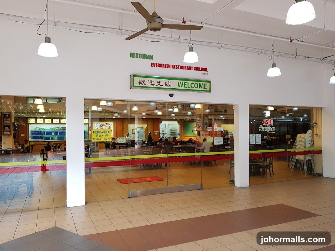 Glass storefront with red and green signage displaying "RESTORAN EVERGREEN RESTAURANT SDN.BHD." and "歡迎光臨 WELCOME" above the entrance. Interior visible through glass doors showing dining area, tables, chairs, and patrons. Ceiling fans and hanging lights above. Yellow and red striped floor mat at entrance. Business hours sign visible inside: 10:30 AM - 10:30 PM. No menu or printed materials clearly visible on walls or counters.