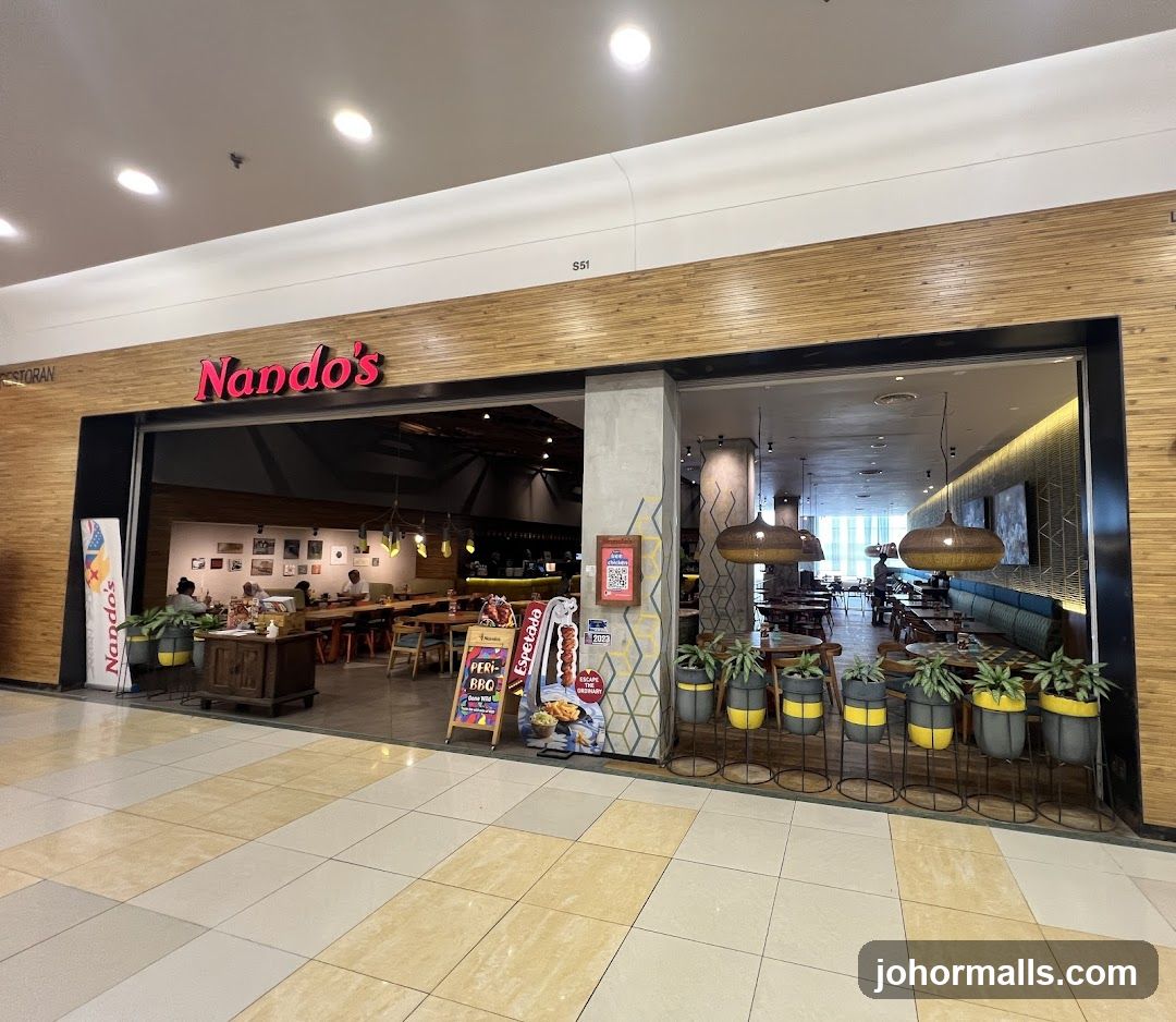 The storefront features a wooden facade with the red 'Nando's' logo prominently displayed above the entrance. The interior is visible through large glass windows, showing dining tables, hanging lamps, and potted plants in gray and yellow pots. A QR code sign and promotional stand are near the entrance.