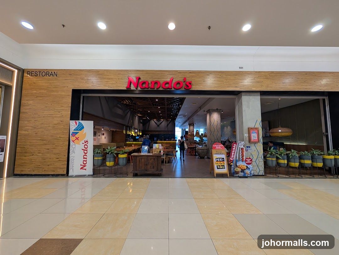 A storefront with a wooden facade featuring the red 'Nando's' logo above the entrance. The interior is visible through the open doorway, showing dining tables, hanging lights, and potted plants in yellow-and-gray pots. A sign on the left reads 'RESTORAN'. Promotional materials like flyers and branded merchandise are placed near the entrance.