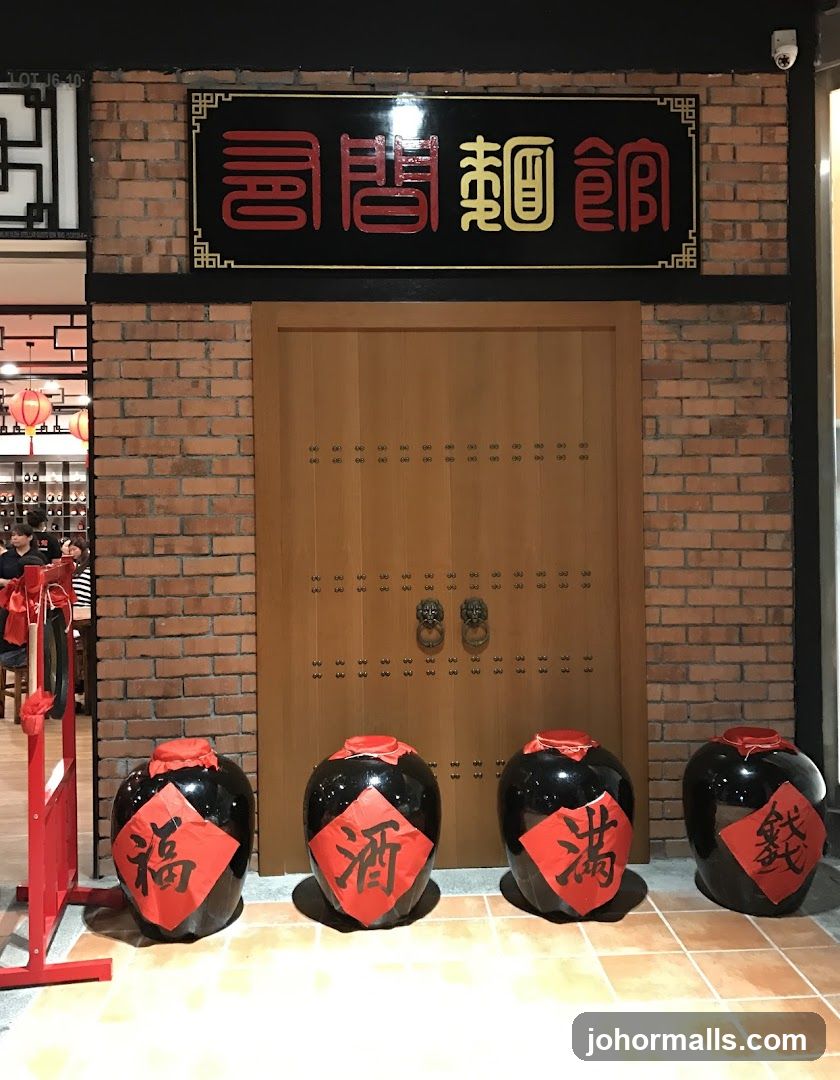 A brick wall facade with a large wooden door. Above the door is a black sign with red and yellow Chinese characters. Four large black ceramic jars with red paper decorations are placed in front of the door. To the left, a red metal barrier and a glimpse of the restaurant interior with tables and chairs are visible.