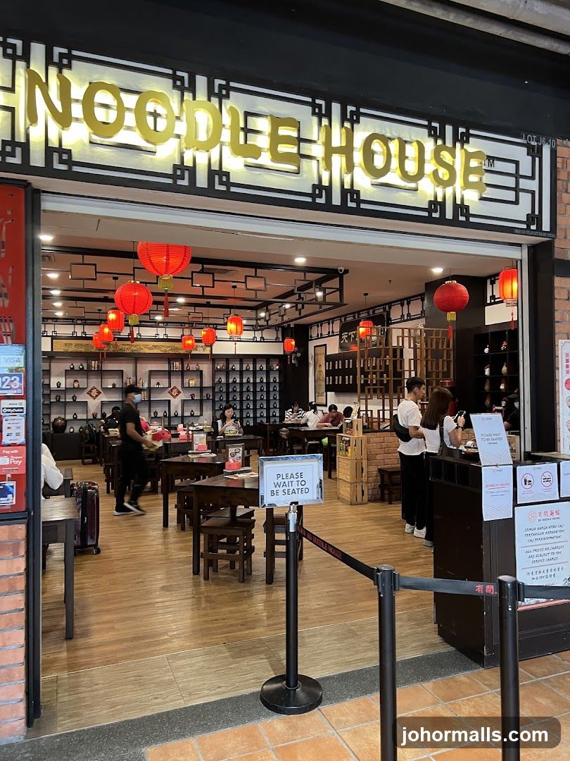The exterior of a restaurant called Noodle House. The entrance is open, revealing tables and chairs inside. Red lanterns hang from the ceiling. Several people are seated at tables, and staff members are visible. A sign at the entrance reads "PLEASE WAIT TO BE SEATED."