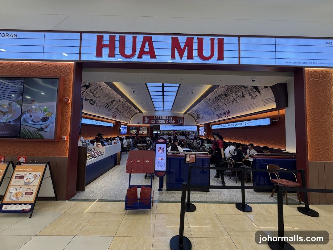 The exterior of the Hua Mui restaurant, with its prominent red lettering signage above the entrance. The restaurant interior is visible, showing seating areas and staff. A digital display on the left shows food images. A-frame signs with menu details are placed outside the entrance.