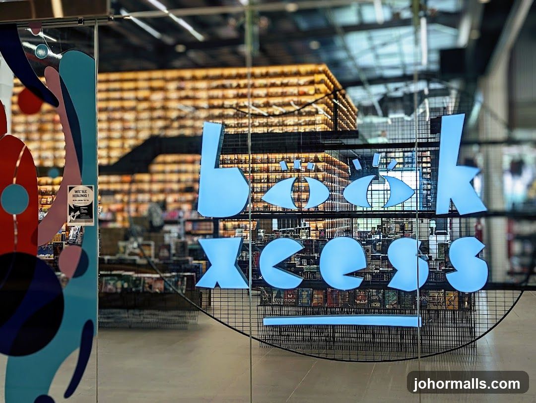 A view through a glass door into a bookstore. The store's name, "Book Access," is displayed in large, light blue, stylized letters on the glass. The letters are designed to resemble eyes and Pac-Man characters. Behind the lettering, shelves filled with books are visible, illuminated by warm lighting. A colorful abstract design is on the left side of the glass. A "We're Hiring" sign is also visible on the door.