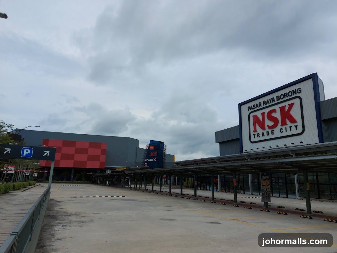 A wide shot of the exterior of NSK Trade City, a large wholesale market. The prominent sign for NSK TRADE CITY is visible, along with "PASAR RAYA BORONG" above it. To the left, there are signs indicating parking and directions. The building features a covered walkway in front of the shops.
