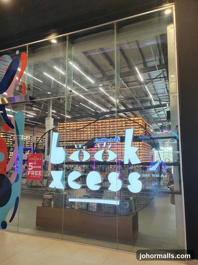 A view through a glass storefront into a bookstore. The store name "book excess" is displayed in large, illuminated white letters on the glass. Inside, shelves filled with books are visible, along with lighting and ceiling structures. A promotional sign on the left reads "BUY 4 BOOKS & GET 5TH BOOK FREE".