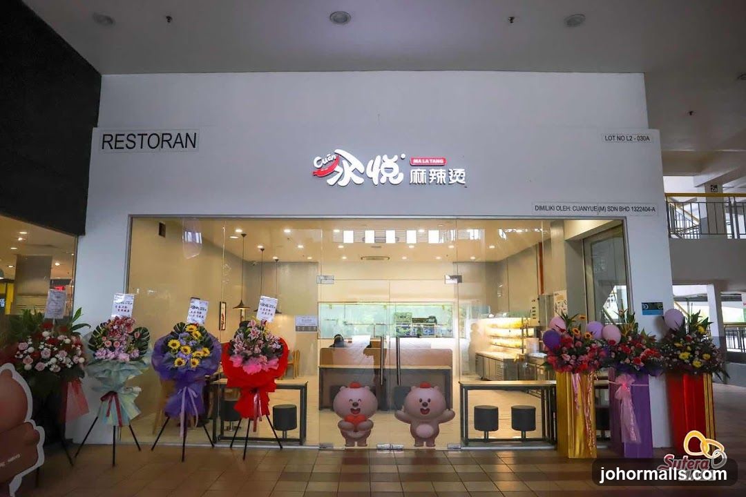 The exterior of a restaurant with a large glass storefront. The restaurant's name, "Cuar Mala Tang" (永悦麻辣烫), is displayed prominently above the entrance. Several congratulatory flower arrangements are placed outside the entrance. Two cartoon bear figures are visible on the glass doors.