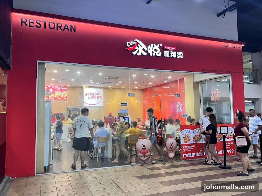 The exterior of a brightly lit restaurant with a red facade and large glass windows. The restaurant's name, "RESTORAN CITAN MA LA TANG" and Chinese characters are visible above the entrance. Inside, customers are seated at tables, and staff can be seen. Decorative cartoon characters are placed near the entrance. Promotional posters are visible on the glass and inside.
