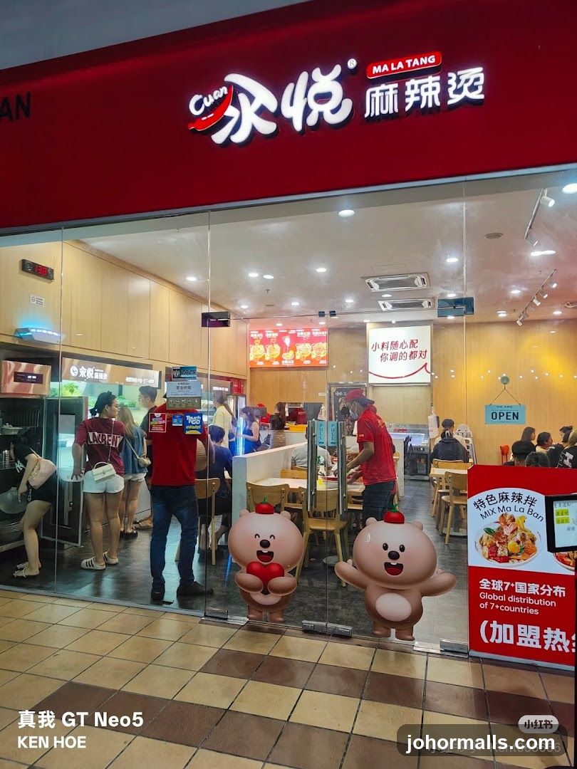 The exterior of a Cuan Ma La Tang restaurant with a red awning and large glass windows. The restaurant's logo is visible above the entrance. Inside, customers are seated at tables, and staff members are visible. Two cartoon bear figures are placed on the glass doors.