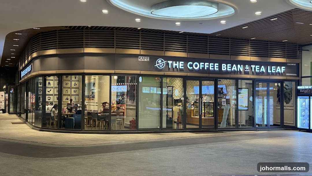 The exterior of a "The Coffee Bean & Tea Leaf" cafe. The storefront features large glass windows and doors, with the brand's name prominently displayed in illuminated lettering above the entrance. Inside, customers can be seen seated at tables, and displays of food and drinks are visible. The cafe has a modern design with a dark facade and warm interior lighting.