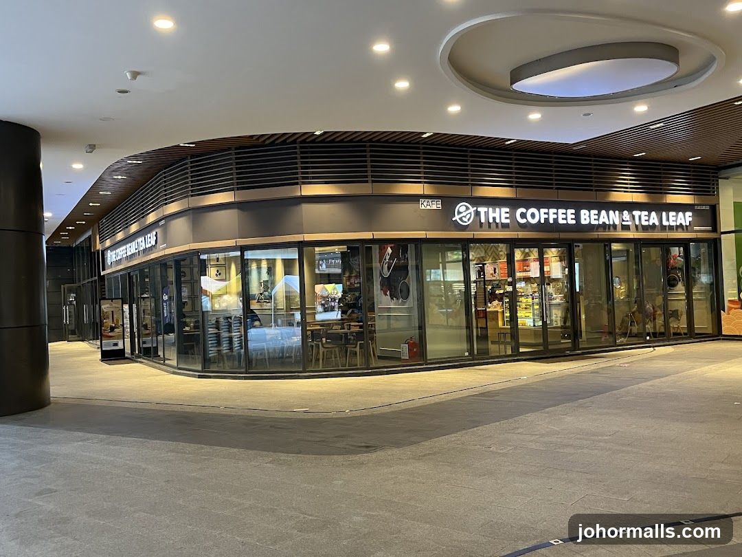 The exterior of a Coffee Bean & Tea Leaf cafe. The storefront features large glass windows and doors, with the cafe's name prominently displayed in illuminated lettering above the entrance. Inside, tables and chairs are visible, along with displays of food and beverages. A sign on an easel stands outside the entrance.